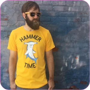 Bearded man wearing the Hammer Time Unisex Adult Tee and bamboo sunglasses, standing in front of a blue brick wall