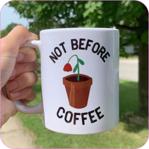 Rhonda's hand holding the Not Before Coffee Mug on a bright, sunny day, with grass and a tree in the background